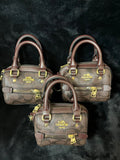 Three Coach mini satchel handbags in brown signature monogram canvas with dark brown leather handles and gold-tone hardware, arranged on a black textured background.